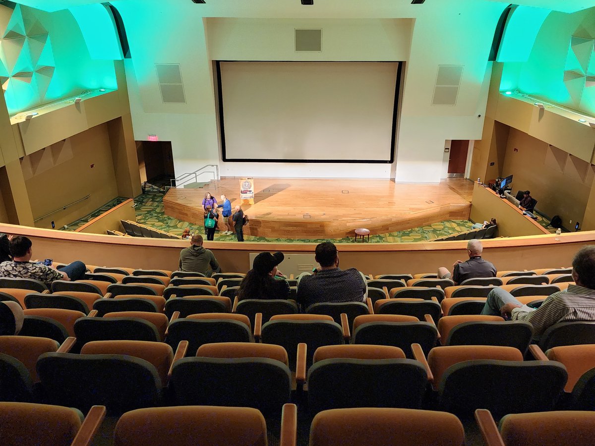 The main auditorium at BSides Tampa: rows of cushioned seats facing a stage with a large projector screen, a few attendees scattered in the seats, teal accent lighting on the side walls.