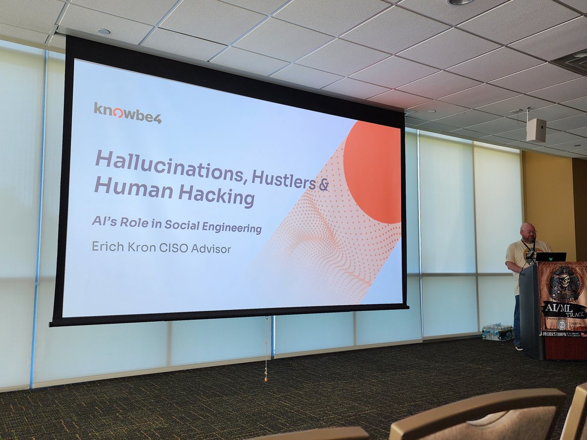 A projector screen at BSides Tampa showing a slide titled 'Hallucinations, Hustlers and Human Hacking — AI's Role in Social Engineering' by Erich Kron, CISO Advisor at KnowBe4. A speaker stands at a podium on the right.
